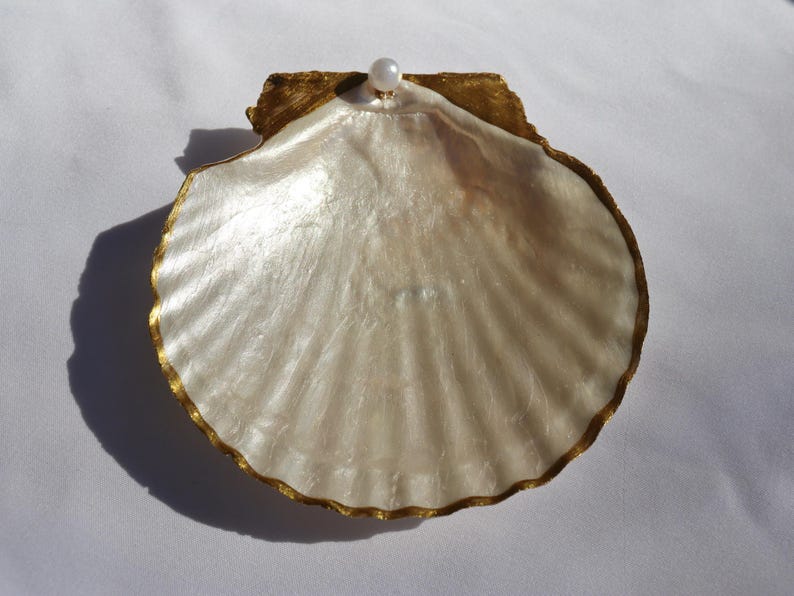 Puede incluir: Una concha de vieira decorativa y abierta con un acabado nacarado. Los bordes de la concha est&aacute;n adornados con oro y una peque&ntilde;a perla se encuentra en la parte superior. La concha tiene una forma natural y el efecto general es elegante y costero.