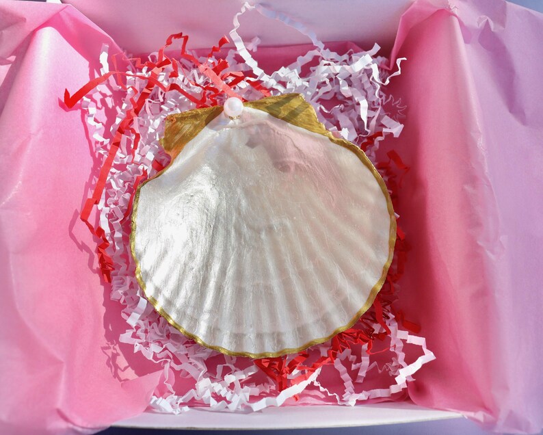 Puede incluir: Una gran concha blanca nacarada con un borde dorado y un peque&ntilde;o acento de perla. La concha est&aacute; colocada en una caja rosa con relleno de papel rojo y blanco, creando una presentaci&oacute;n de regalo.