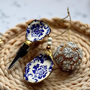 May include: A decorative wine bottle stopper and two oyster shell ornaments with blue and white floral designs and gold trim. A beaded ornament and a woven coaster are also in the image.