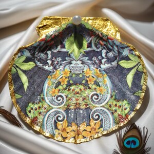 May include: Decorative seashell with a gold-leaf edge and a pearl accent. The interior features a detailed floral and scroll design in shades of blue, green, and yellow. The shell rests on a white satin surface.