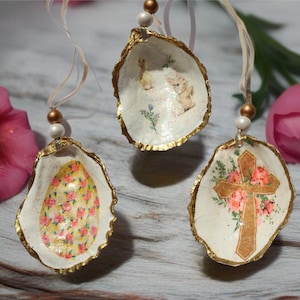 May include: Three decorative oyster shell ornaments with gold edges. One features a floral egg design, another has a cross with flowers, and the third displays bunnies. Each has a beaded string for hanging, perfect for Easter.