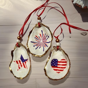 Puede incluir: Tres adornos de concha de ostra con diseños patrióticos. Cada concha tiene un borde dorado y está decorada con imágenes rojas, blancas y azules, incluyendo una estrella, fuegos artificiales y una bandera americana en forma de corazón. Se adjuntan cintas rojas para colgar.