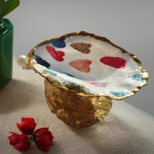 Puede incluir: Un cuenco decorativo de concha de ostra con exterior y borde pintados en dorado. El interior es blanco con patrones de corazones coloridos. Una perla está unida al borde. El cuenco se muestra con un jarrón verde y flores rojas.