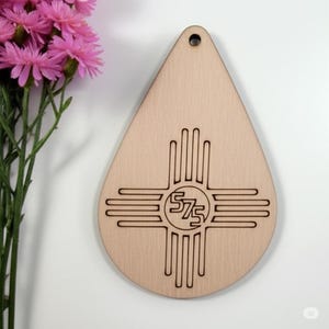 May include: A wooden teardrop-shaped ornament with a laser-etched design. The design features a stylized sun symbol with the numbers "575" in the center. The ornament has a small hole at the top for hanging. Pink flowers are in the background.