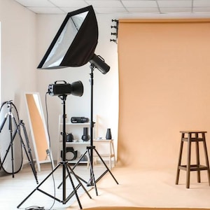 Op de afbeelding: Een fotostudio-opstelling met verlichtingsapparatuur, een achtergrond en een houten kruk. De studio beschikt over softboxen, lampen op statieven en een neutrale achtergrond.