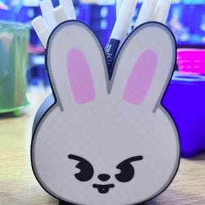 May include: A white and black bunny-shaped desk organizer holding white markers. The bunny has pink inner ears and a mischievous expression. The organizer is on a wooden surface, with other office supplies in the background.