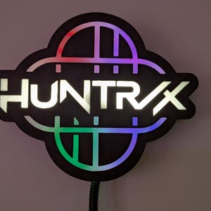 May include: Black sign with the word "HUNTRAX" in white, illuminated letters. The sign has a unique shape with a colorful, glowing border in shades of red, purple, green, and blue. A black cord extends from the bottom.