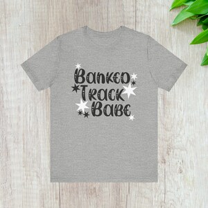 Könnte beinhalten: Ein hellgraues T-Shirt mit dem Text "Banked Track Babe" in schwarzer Schrift, verziert mit weißen Sternen. Das Shirt wird auf einer hellen Holzoberfläche präsentiert.