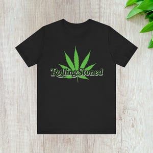 May include: Black t-shirt with a green cannabis leaf graphic and the text "Rolling Stoned". The design is centered on the front. The shirt is made of a soft material. The text and leaf are in a contrasting green color.