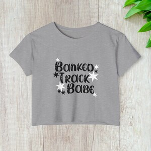 Puede incluir: Un top corto gris con el texto "Banked Track Babe" en escritura negra y estrellas. La camiseta está hecha de un material suave y es adecuada para uso casual.