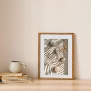 May include: A framed print with a zen garden design featuring smooth stones and raked sand patterns. The artwork is in a wooden frame and is displayed on a wooden surface with a stack of books and a ceramic mug.