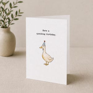 Könnte beinhalten: Eine weiße Grußkarte mit einer Aquarellillustration einer hellbraunen Ente, die einen kleinen blauen Partyhut mit einem roten Bommel trägt. Der Text, der in einer Schreibmaschinenschrift über der Ente gedruckt ist, lautet: Have a quacking birthday.