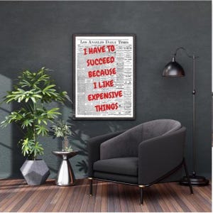 Könnte beinhalten: Ein gerahmter Druck mit dem Text "I HAVE TO SUCCEED BECAUSE I LIKE EXPENSIVE THINGS" in Rot auf einem Zeitungs-Hintergrund. Der Druck hängt an einer dunklen Wand über einem schwarzen Sessel. Eine Stehlampe und eine Topfpflanze sind ebenfalls zu sehen.