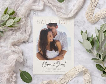May include: A cream-colored wedding invitation card with the text "Save the Date" at the top. The card features a photo of a couple embracing. The names "Olivia & Daniel" and the date "14 October 2022" are written below the photo. The card is surrounded by decorative elements.