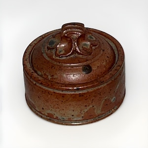 May include: A circular, lidded ceramic pot in shades of brown. The lid has a floral design and a small handle. The pot has a rustic, speckled appearance, suggesting a handmade or vintage origin. The pot is likely used for storage, with a diameter of approximately 10 cm.