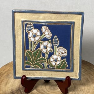 May include: Square ceramic tile with a floral design. The tile features white flowers with green leaves against a blue background, framed by a beige border with a blue trim. The tile is displayed on a wooden stand.