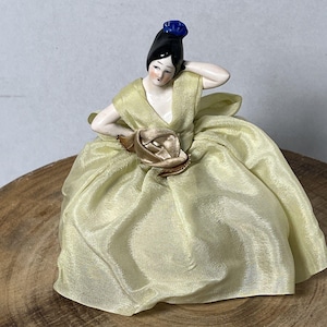 Può includere: Statuina vintage in porcellana di una donna in un abito giallo pallido e fluente. La statuina ha capelli neri, un copricapo blu e un accento di rosa dorata. La figura è in posa con un braccio alzato.