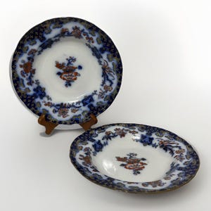May include: Two antique porcelain plates with a white center and a blue floral border. The plates feature a central floral design in orange and red. One plate is on a wooden stand.