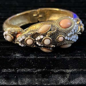 May include: Gold-tone hinged bracelet with a textured design. The bracelet is adorned with peach, cream, and white-colored stones. The stones are round, oval, and teardrop-shaped, adding a touch of elegance to the jewelry.