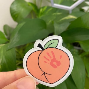 May include: A peach-shaped sticker with a white border. The peach is a light orange color with a red handprint on one side. A green leaf and black stem are at the top. The sticker is held in front of a background of green leaves.