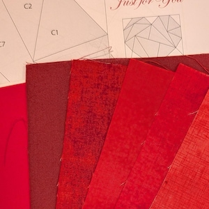 May include: A set of six fabric swatches in varying shades of red, ranging from deep burgundy to bright orange-red, arranged in a fan shape. Behind the fabric is a paper pattern labeled Quilt Block Just for You with geometric shapes.