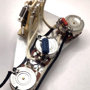 May include: Close-up of a guitar control plate with various electronic components. The plate is silver and holds potentiometers, a switch, and wiring. The components are connected with white and black wires, and a blue capacitor is visible.