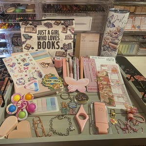 May include: A collection of stationery and accessories, including stickers, pens, bookmarks, and a tumbler, arranged on a gray tray. The items are in shades of pink, peach, and pastel colors. A canvas bag with the text "JUST A GIRL WHO LOVES BOOKS" is also visible.