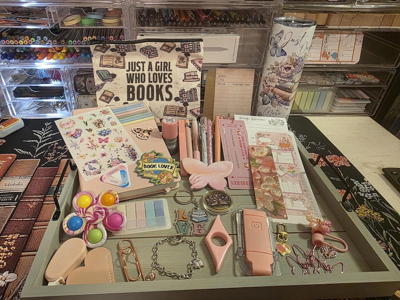 May include: A collection of stationery and accessories, including stickers, pens, bookmarks, and a tumbler, arranged on a gray tray. The items are in shades of pink, peach, and pastel colors. A canvas bag with the text "JUST A GIRL WHO LOVES BOOKS" is also visible.