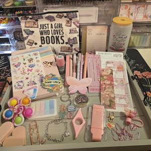 May include: A collection of stationery and accessories, including notebooks, pens, stickers, and a pencil case with the text "JUST A GIRL WHO LOVES BOOKS." The items are arranged on a tray, with a color palette of pink, pastel colors, and gold accents.