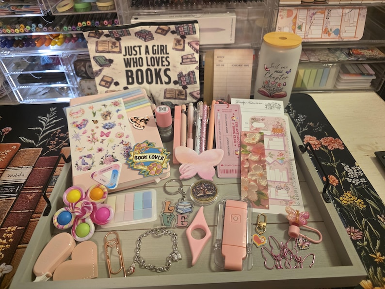 May include: A collection of stationery and accessories, including notebooks, pens, stickers, and a pencil case with the text "JUST A GIRL WHO LOVES BOOKS." The items are arranged on a tray, with a color palette of pink, pastel colors, and gold accents.