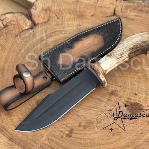 May include: A black blade knife with a deer antler handle and a leather sheath. The sheath is embossed with a leaf design. The knife is lying on a wooden surface. The text "Sh Damascus" is visible.