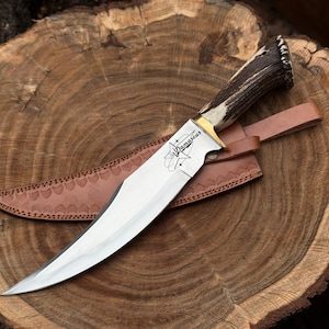 May include: A Damascus steel knife with a polished blade and a handle made of antler material. The knife rests on a brown leather sheath, with the word "Damascus" etched on the blade. The knife is displayed on a wooden surface.
