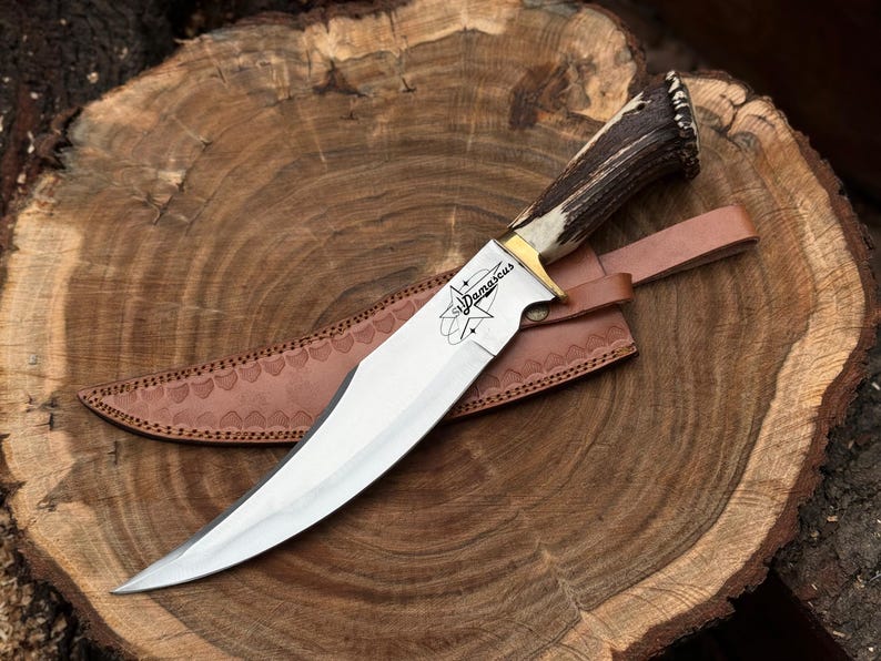 May include: A Damascus steel knife with a polished blade and a handle made of antler material. The knife rests on a brown leather sheath, with the word "Damascus" etched on the blade. The knife is displayed on a wooden surface.