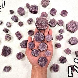 May include: A collection of raw, hexagonal-shaped purple and magenta corundum crystals scattered across a surface, with several pieces resting in an open palm. The stones feature a rough, matte texture with visible crystalline structures throughout.