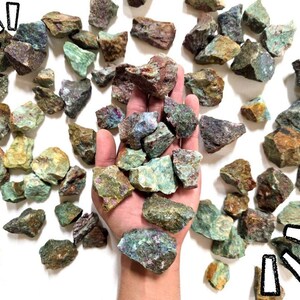 May include: A collection of raw, unpolished mineral stones featuring a mix of green, brown, and reddish-purple hues. The stones have irregular, jagged shapes and varied textures, with some pieces displaying metallic or crystalline surfaces throughout.