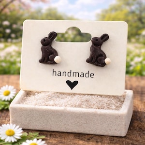 May include: A pair of dark brown bunny earrings with white tail accents are displayed on a white card that reads "handmade" with a heart symbol. The card is set in a small, rectangular, sand-filled container, with daisies in the foreground.