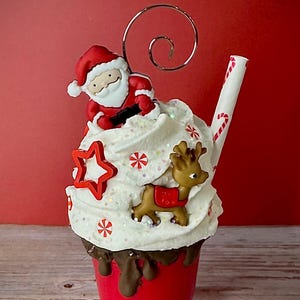 May include: A Christmas decoration in the shape of a hot chocolate cup. It has a red cup with whipped cream, a Santa Claus figure, a reindeer, and a candy cane straw. The whipped cream is decorated with red star and snowflake accents.