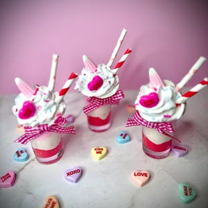 May include: Three Valentine's Day desserts in clear glasses, each with whipped cream, pink heart-shaped candies, and unicorn horn decorations. Red and white striped straws and pink gingham ribbons enhance the festive theme. Conversation heart sweets are scattered around.