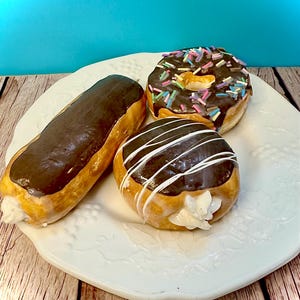 May include: A white plate holds three glazed pastries. An éclair with chocolate frosting and cream filling sits next to a round donut with chocolate frosting and sprinkles. A second donut has white icing stripes and cream filling.