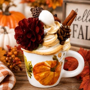 May include: A white ceramic mug decorated with a painted orange pumpkin, topped with a decorative arrangement of a large dark red pinecone, a white egg-shaped ornament, a small brown pinecone, and a cinnamon stick, resembling a festive drink.