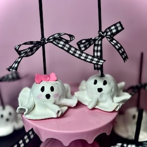 May include: Two white ghost-shaped treats with black eyes and mouths, perched on a pink cake stand. Each ghost has a black stick and a black and white checkered bow. One ghost has a pink bow on its head. The background is pink.