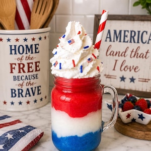 May include: A glass mason jar mug filled with a layered red, white, and blue frozen beverage topped with a large swirl of whipped cream, small American flag sprinkles, and a red and white striped paper straw, creating a patriotic drink.