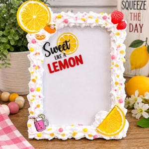 May include: A white picture frame decorated with whipped cream-like frosting, yellow lemon slices, and pink sprinkles. The frame has the words "Sweet like a Lemon" in black and red. A small pink drink with a straw is on the bottom left.