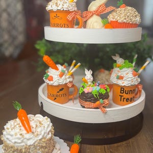 May include: A two-tiered white serving tray with Easter-themed decorations. The tray holds orange mugs with the word "Carrots" and "Bunn Treat", cupcakes, a bunny, and carrot decorations. The items are decorated with white frosting and orange accents.