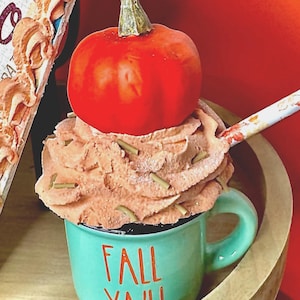 May include: A teal mug with the words "FALL YA'LL" in red lettering, filled with a brown, whipped topping and topped with a small orange pumpkin. A spoon with a floral design rests on the topping. The mug sits in a wooden bowl.