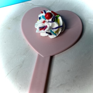 May include: A pink heart-shaped hand mirror topped with a dollop of white frosting. The frosting is decorated with colorful sprinkles and a red cherry. The mirror's handle extends downward.