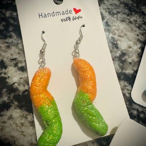 May include: A pair of novelty earrings shaped like orange and green gummy worms. The earrings are attached to silver-colored fish hook ear wires. The earrings are displayed on a white card with the text "Handmade with love".