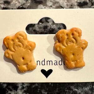 May include: A pair of handmade, light brown bear-shaped earrings. The earrings are displayed on a white card with a black heart and the word "ndmade". The earrings are made of a light brown material.