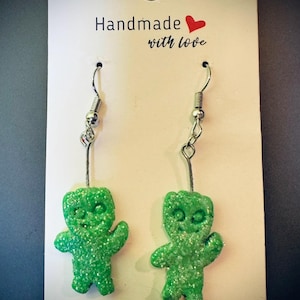 May include: A pair of handmade green gummy bear earrings with silver-colored hooks. The earrings are glittery and have a smiling face. The backing card reads "Handmade with love."