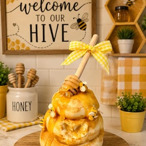 May include: A tiered stack of round, light-colored baked goods topped with a thick, golden honey glaze and small decorative bees. A wooden honey dipper is inserted into the top, adorned with a yellow and white checkered fabric bow.